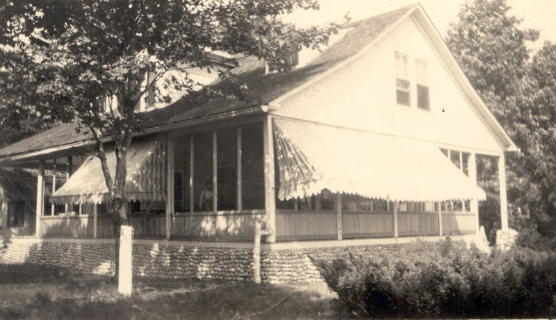 A white summer cottage sits nestled between trees. Behind the low awnings, several people appear on its enclosed porch.