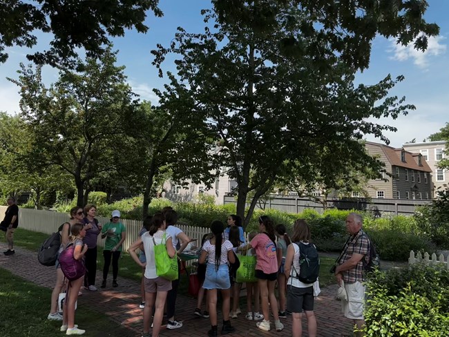A group of people wearing backpacks stand outside surrounding a table. There is a green lush garden behind them with a fence dividing it.
