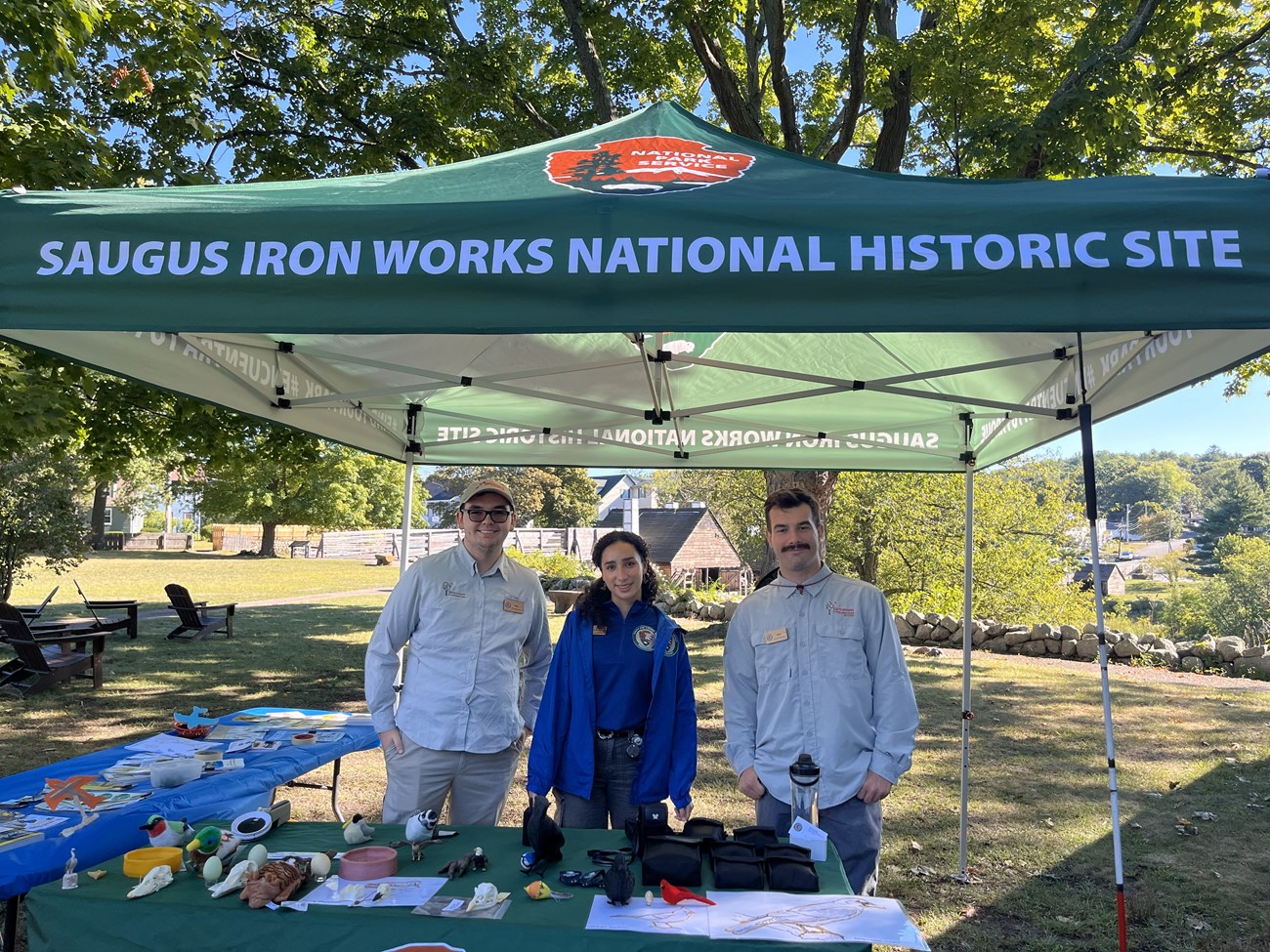 Three young adults stand outside under a green tent that reads “Saugus Iron Works National Historic Site.” They are standing behind a table with paper materials, plastic bird skulls, and egg models.