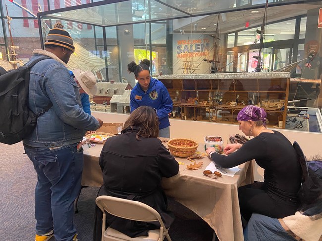 A young woman stands behind a table. In front of her are three young adults standing and sitting around it. They are looking at a collection of things on the table like a basket full of dried milkweed pod.
