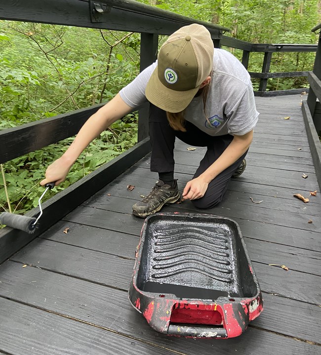 Clara Painting A young woman wearing a green ball cap kneels on a gray wooden bridge while painting the hand rail