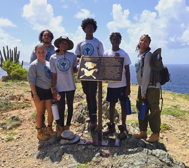A group of six youth stand outside in the dirt standing in front of a sign with water in the background
