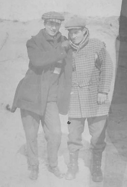 Two men in earm winter clothing stand in front of a sandy dune.