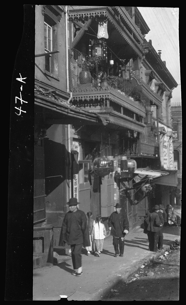 People walk on a sidewalk under balconies hung with lanterns.