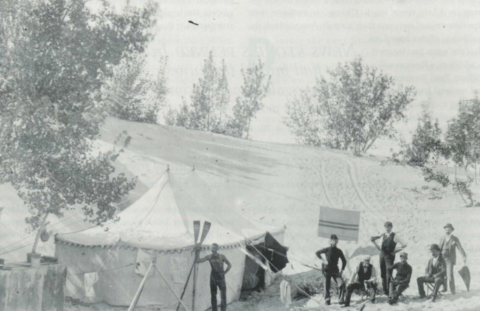 Black and white photograph of seven men, seated and standing near a large white tent in front of a sand dune.