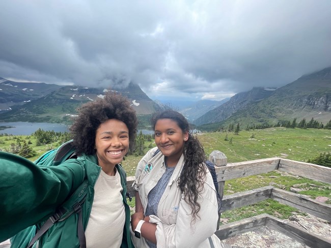 Two young woman smile for a selfie in the outdoors. Vast mountains stand behind them.