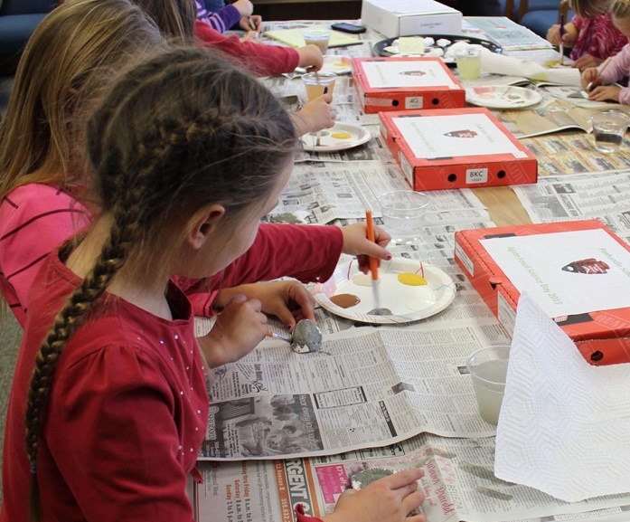 Several young children sit around a table covered with newspapers and paints on paper plates. They paint small figures with paintbrushes.