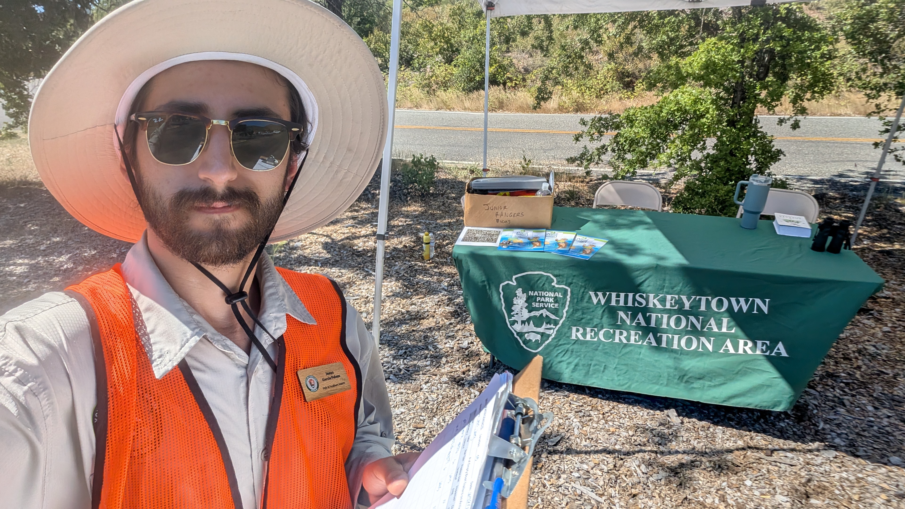 A young man in an orange vest and hat holds a clipboard outside. He is standing in front of a green table that reads “Whiskeytown National Recreation Area.”