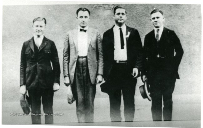 Four men in suits pose in a line holding hats in their right hands.
