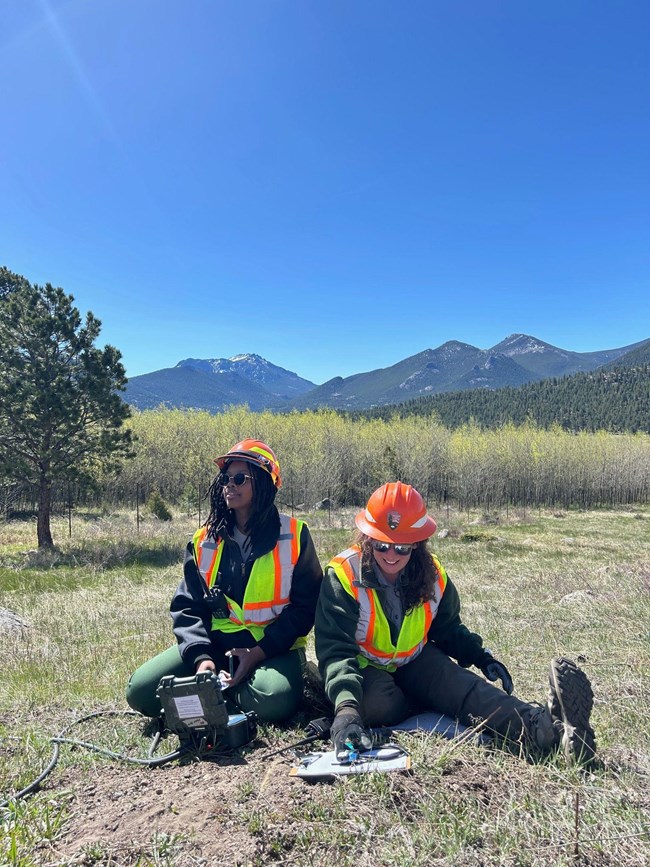 Traffic Monitor Two young women sit in the grass outside wearing orange and green neon vests and hard hats