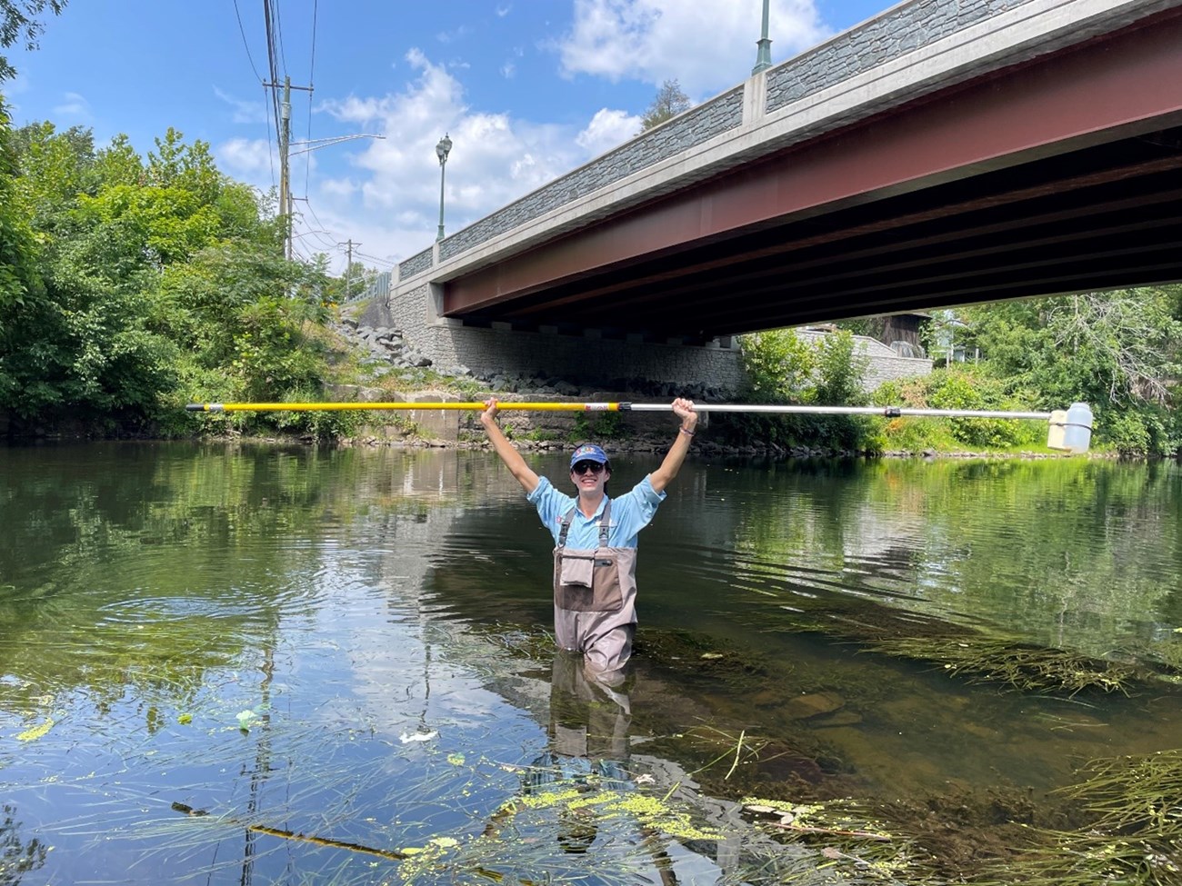 Andres in water A young man stands in a river surrounded by trees. He is holding a yellow pole in the air with both hands and wearing waders.