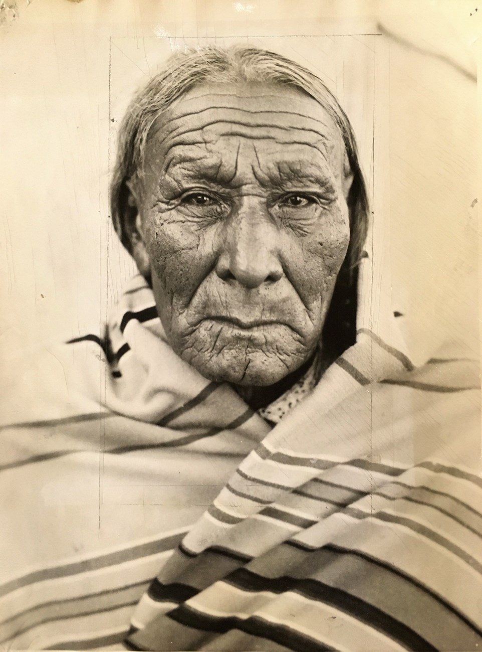 Up close photograph of Chief Magpie's face. He is around age 79. He has wrinkled, tan skin and dark silver hair. He is wrapped in a stripped wool blanket up to his neck.