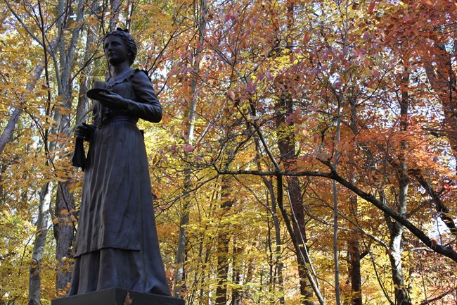 Surrounded by colorful fall foliage, the Turner monument is a statue depicting a woman in a colonial style dress. She holds a saucer and cup in her left hand, and has a towel draped over her right forearm.