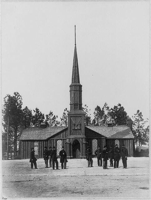 Ten men stand in front of a wooden church with the steeple; Engineering Corp symbol is in the center of the steeple above the door arch.