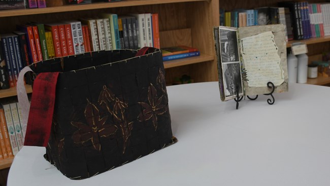 A black basket made from woven paper and red strap in the froeground with a book on a stand in the background.