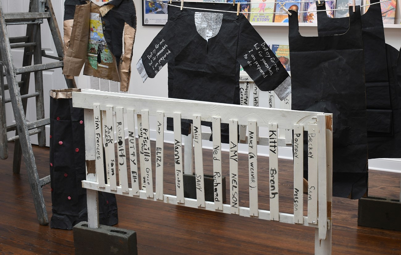 A white picket fence with names painted on the pickets stands in front of black paper garments on a clothesline.