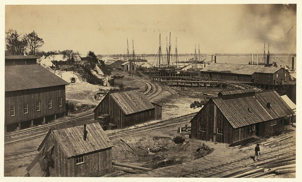 The United States Military Railroad Depot at City Point during the civil war. Mutiple wood buildings and railroad tracks along the edge of the James River.