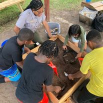 Archeology Activity Four children with brown skin dig in a box of dirt with archeology tools while a black female wearing a NPS uniform supervises.