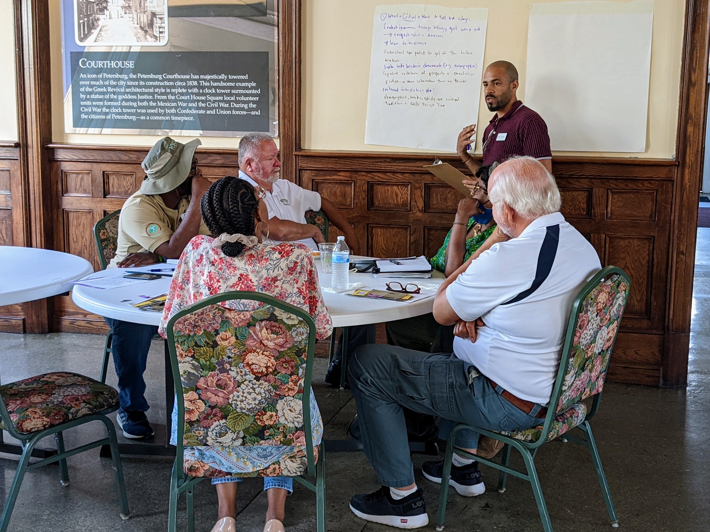 Breakout Session Discussion Five people, black and white, sit at a table, a black male stands near a large white piece of paper taking notes.