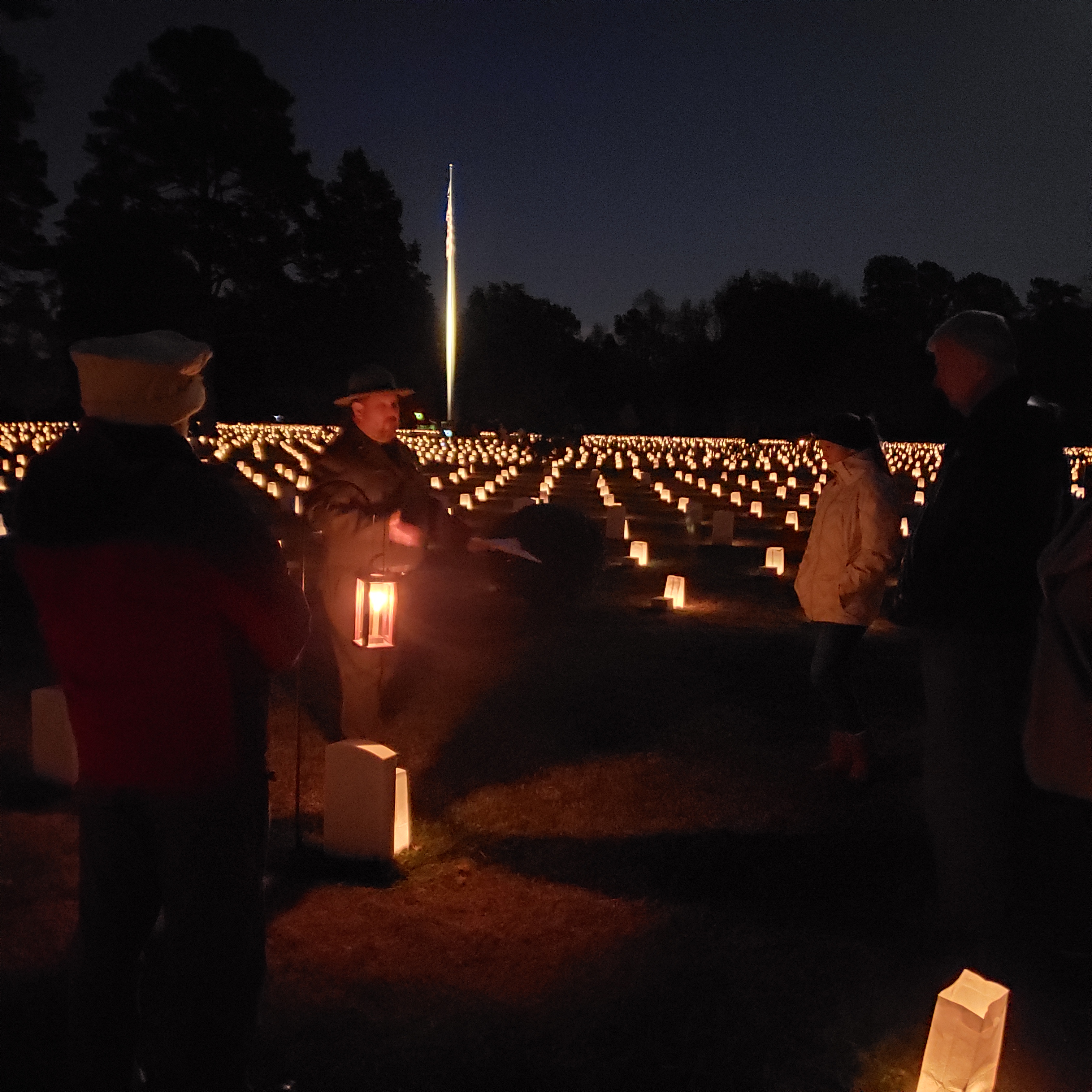 Poplar Grove National Cemetery Luminary Program Dark night-Park Ranger stands next to a lantern on a hook in a cemetery lit by luminaries.