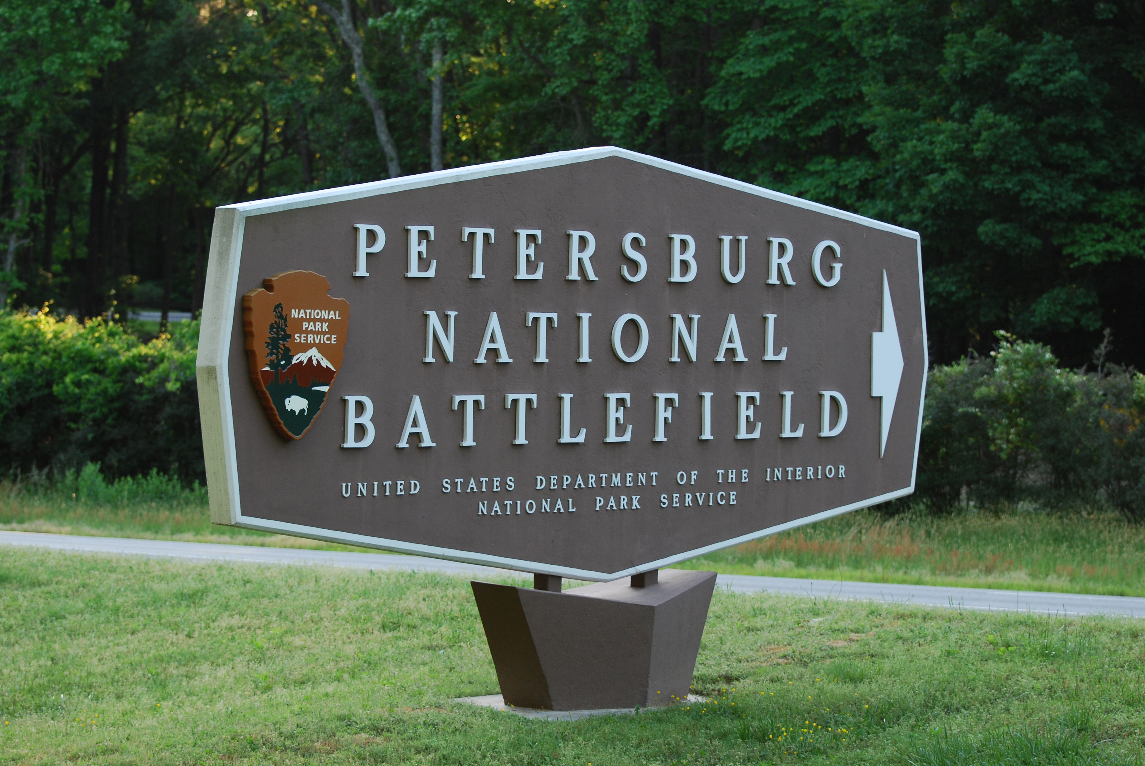 Brown rectangular octagon with white text reading Petersburg National Battlefield; NPS arrowhead on left end of sign and an arrow point right on the right end of the sign.