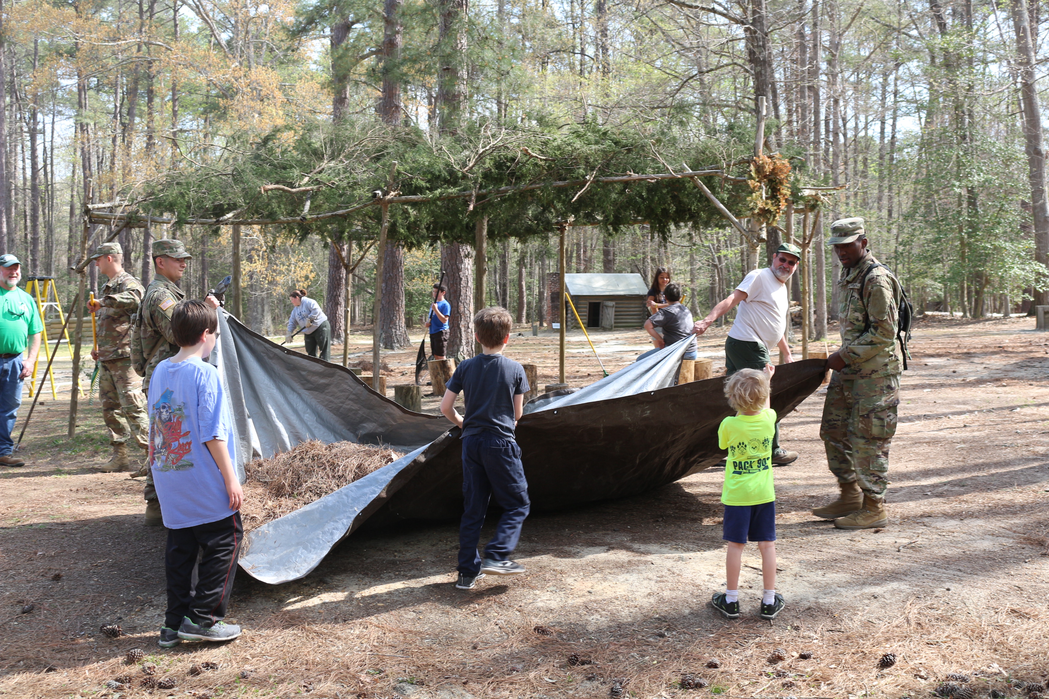 Three children, two soldiers in uniform, and a man drag a trap filled with pine needles. Six other people raking and cleaning in the area. A small cabin and trees are in the background.