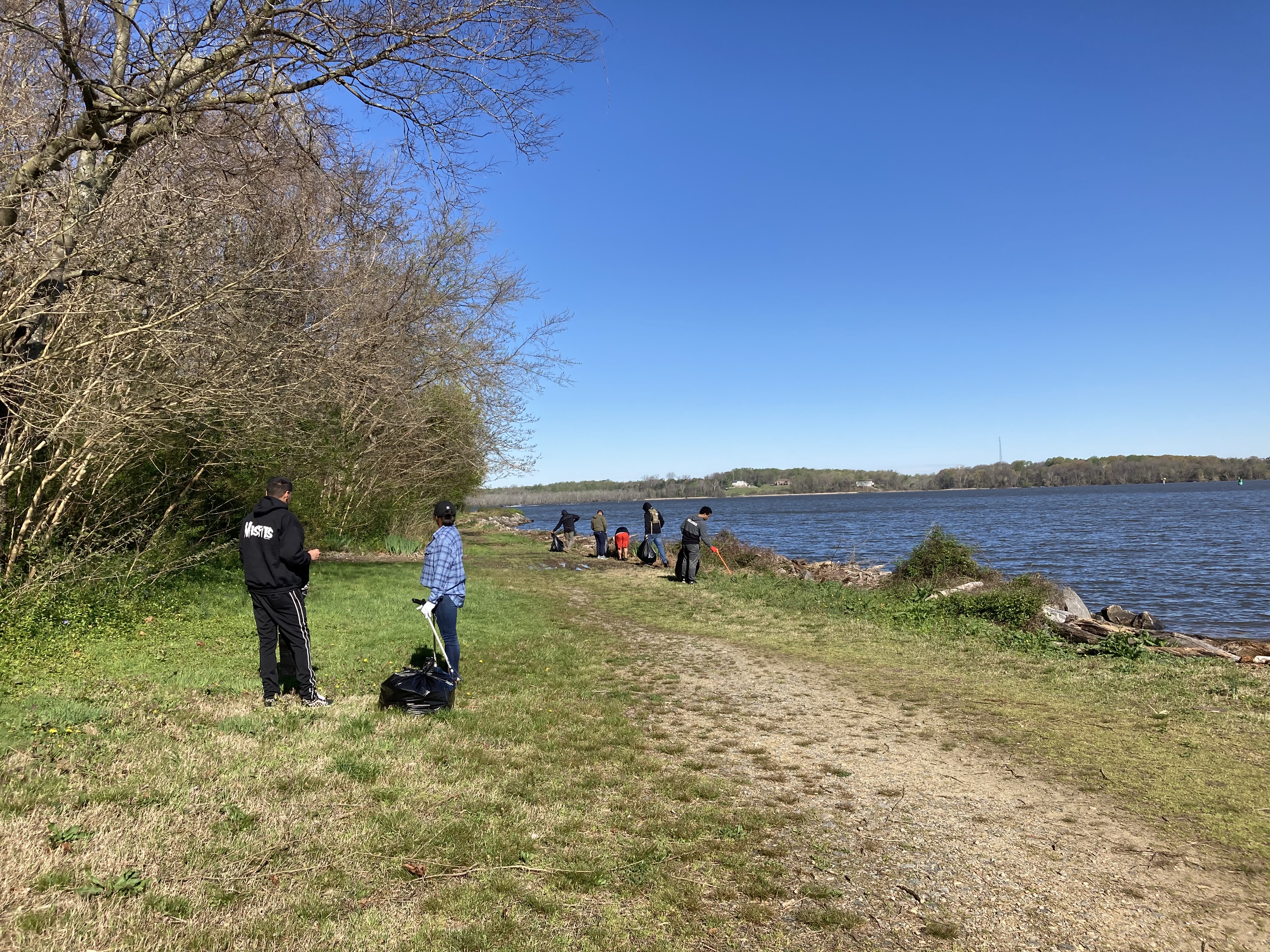 A mixed group of men and women pick up trash on the shorelinr of a river.