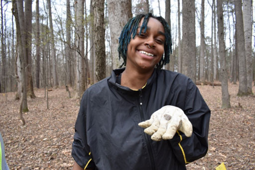 A college aged black woman in the woods. She is holding an arrowhead found in a gloved hand and smiling.