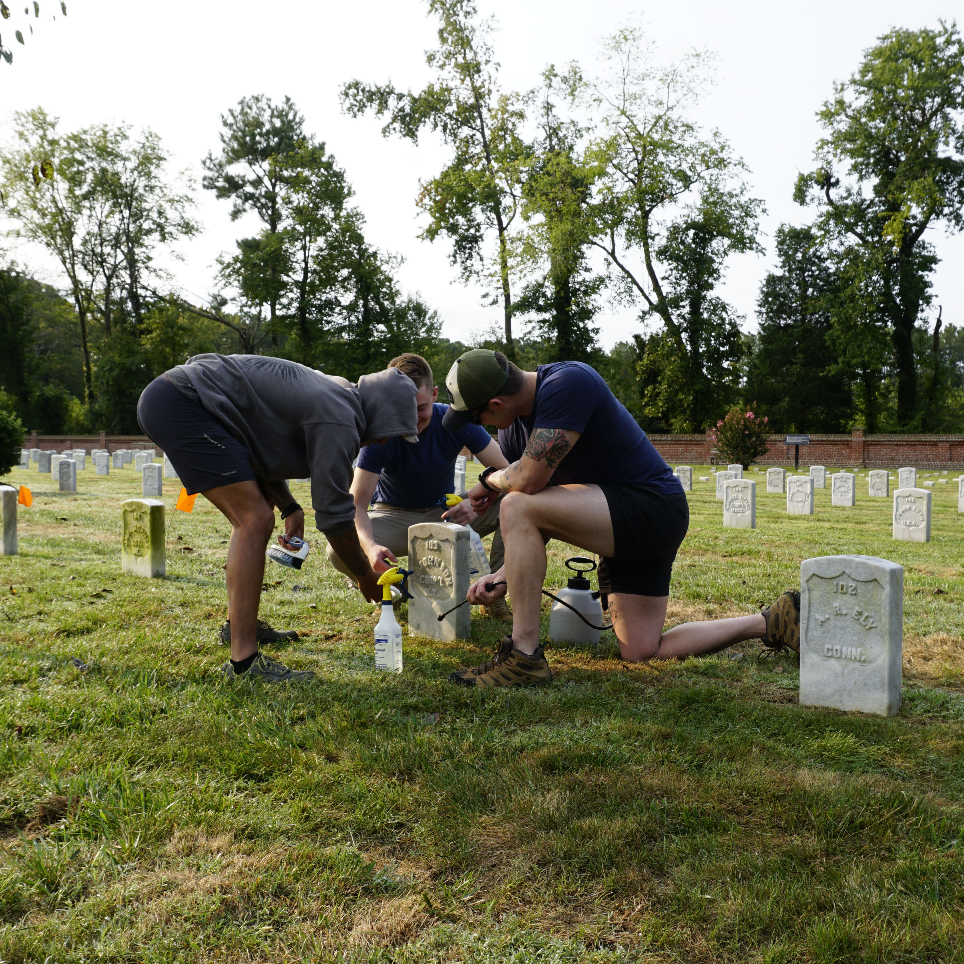 Three people gather around a headstone with cleaning tools.