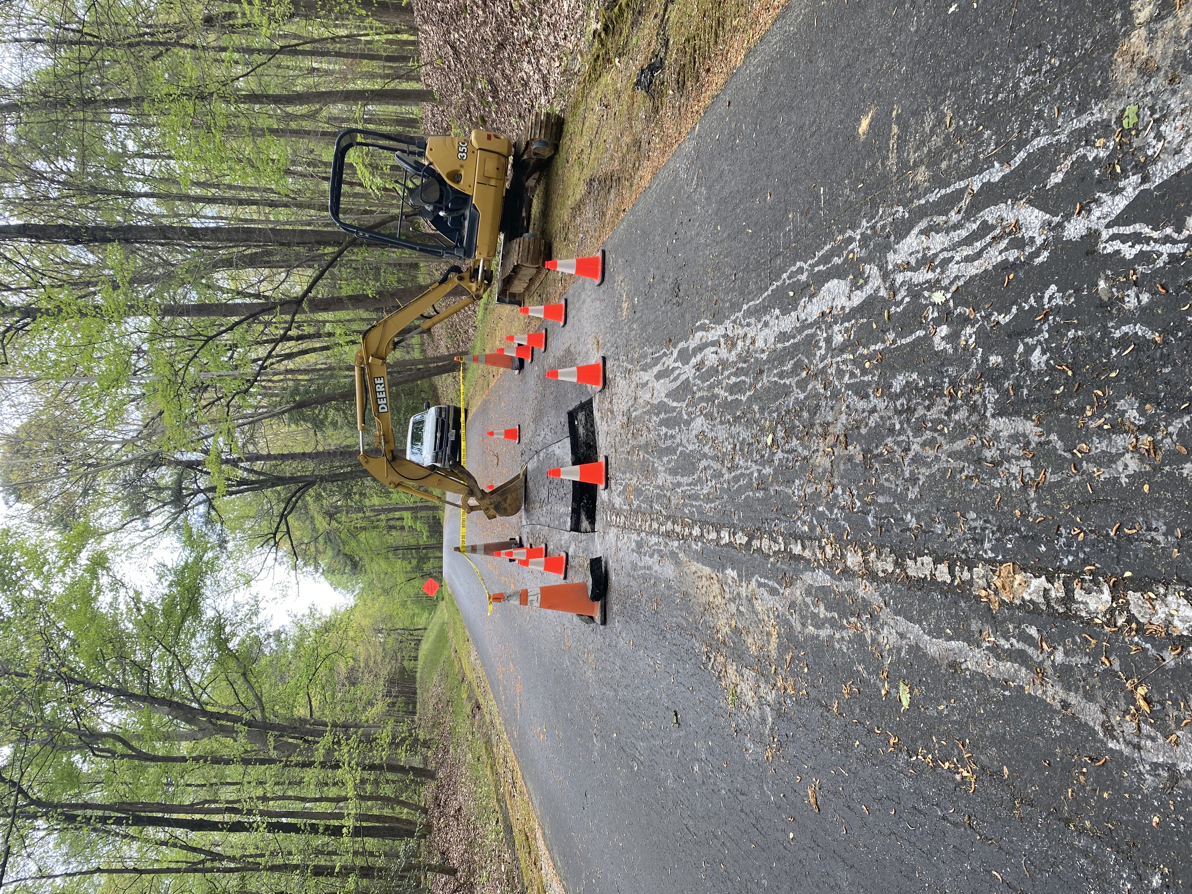 Orange cones surround a hole in a road. A yellow excavator is in the background.