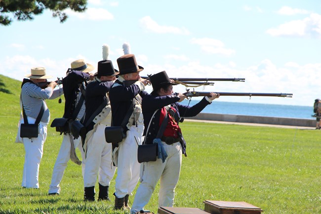Five reenactors in various uniforms in a line at the aim position  with muskets point down range horizontal waiting on the fire command.