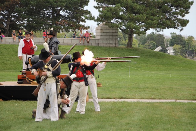 Three reenactors dressed as War of 1812 Infantry and Sailors at different stages of firing their muskets
