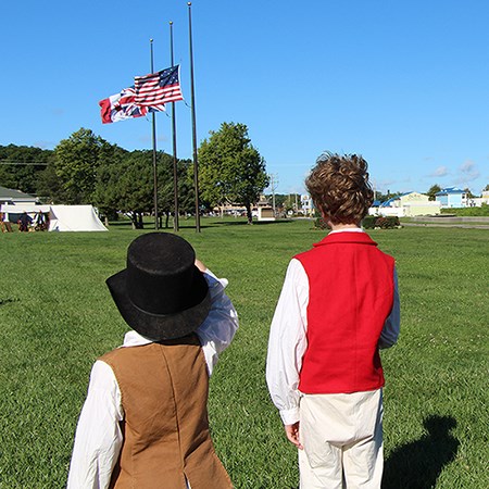 Two young reenactors saluting flags as they are being raised. One dressed as civilian with brown vest and black felt round hat. The other as a sailor with a red vest.