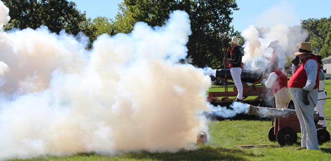 Two pieces of naval artillery fire. Closer is smaller 6 pounder on a naval carriage. Half the picture is smoke filled.