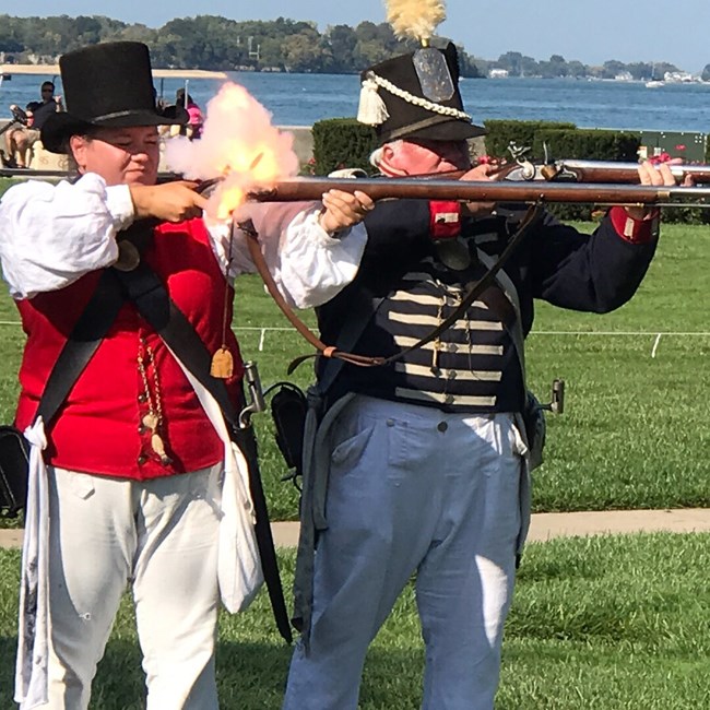 Two people, one dressed as sailor and other infantry from War of 1812 have muskets leveled and pulling trigger. There is a flash of fire and smoke at vent/pan area of musket.