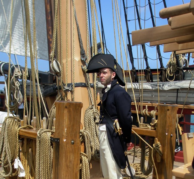 Men dressed as War of 1812 officer. Bicorn hat and dark blue coat with tails stand on the deck of a tall ship near a mast.