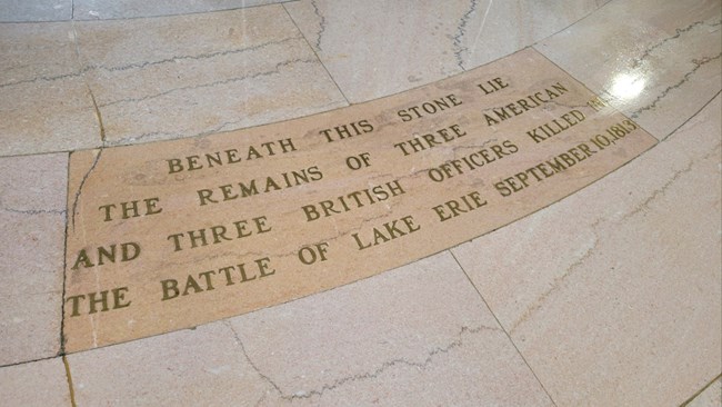 A marble stone with bronze engraving set in a marble floor tiles. It reads "Beneath this stone lie the remains of three American and three British officers killed in the Battle of Lake Erie September 10, 1813.