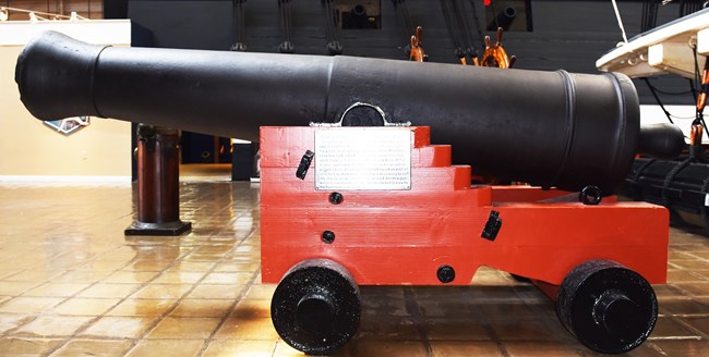 Black cannon tube sits on a red wood naval truck with 4 small black metal wheels.