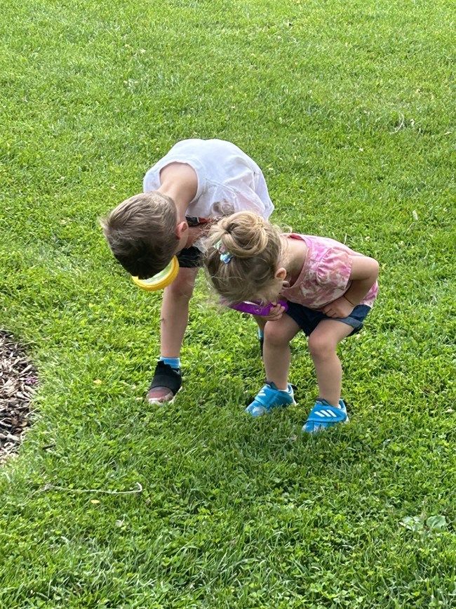 Two young children are bent over, holding and looking through large magnifying glasses at the grass