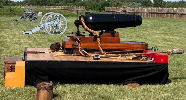 Carronade Short gun tube sits on a wooden skid and sled mount with rope rigging to secure the gun and pull it forward. All of this is mounted on simulated gun deck with a black fabric skirt.