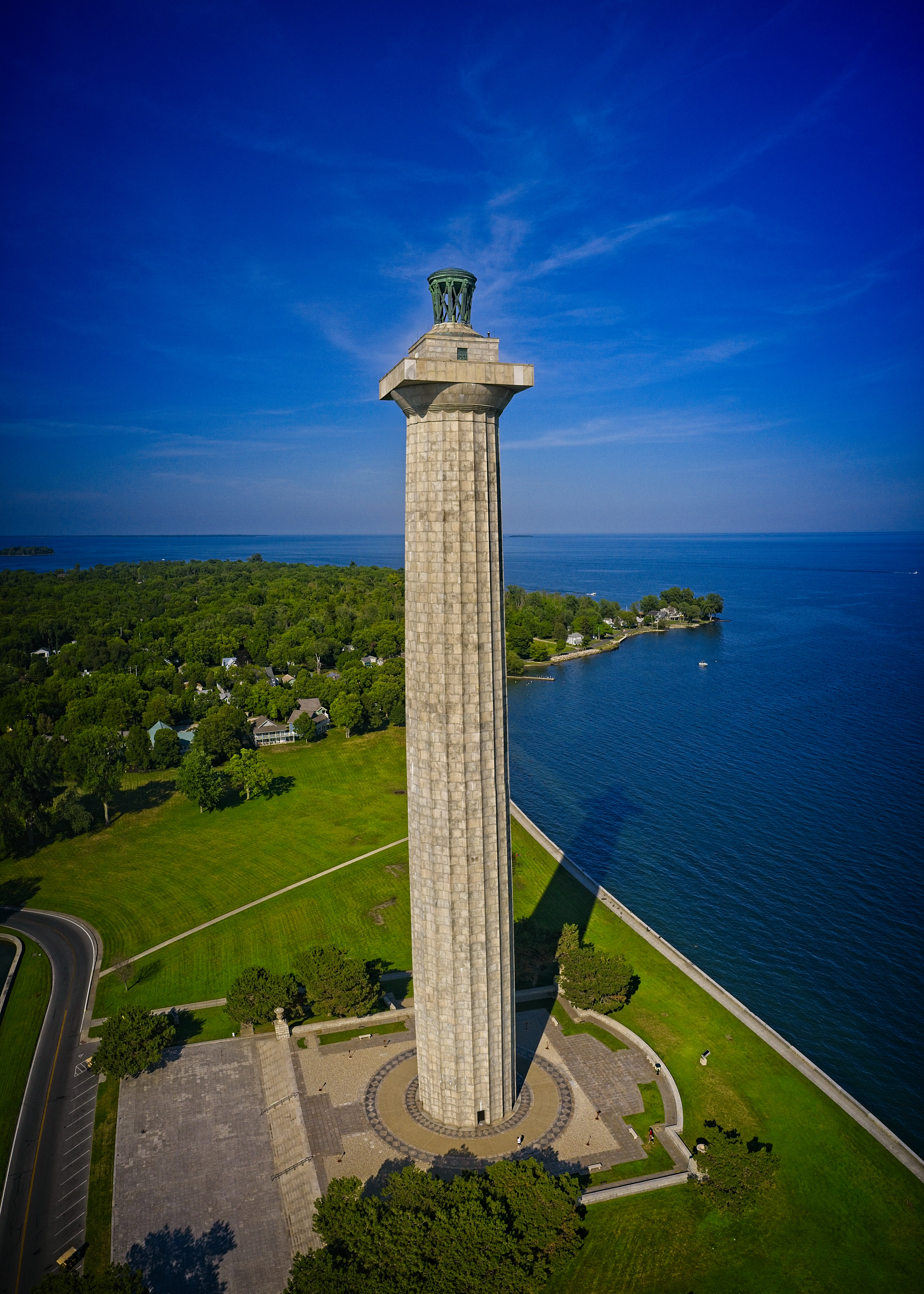 Aerial image of a tall stone memorial with two plazas at base on an isthmus of an island. Surrounded by green grass. Blue sky and water in the distance.