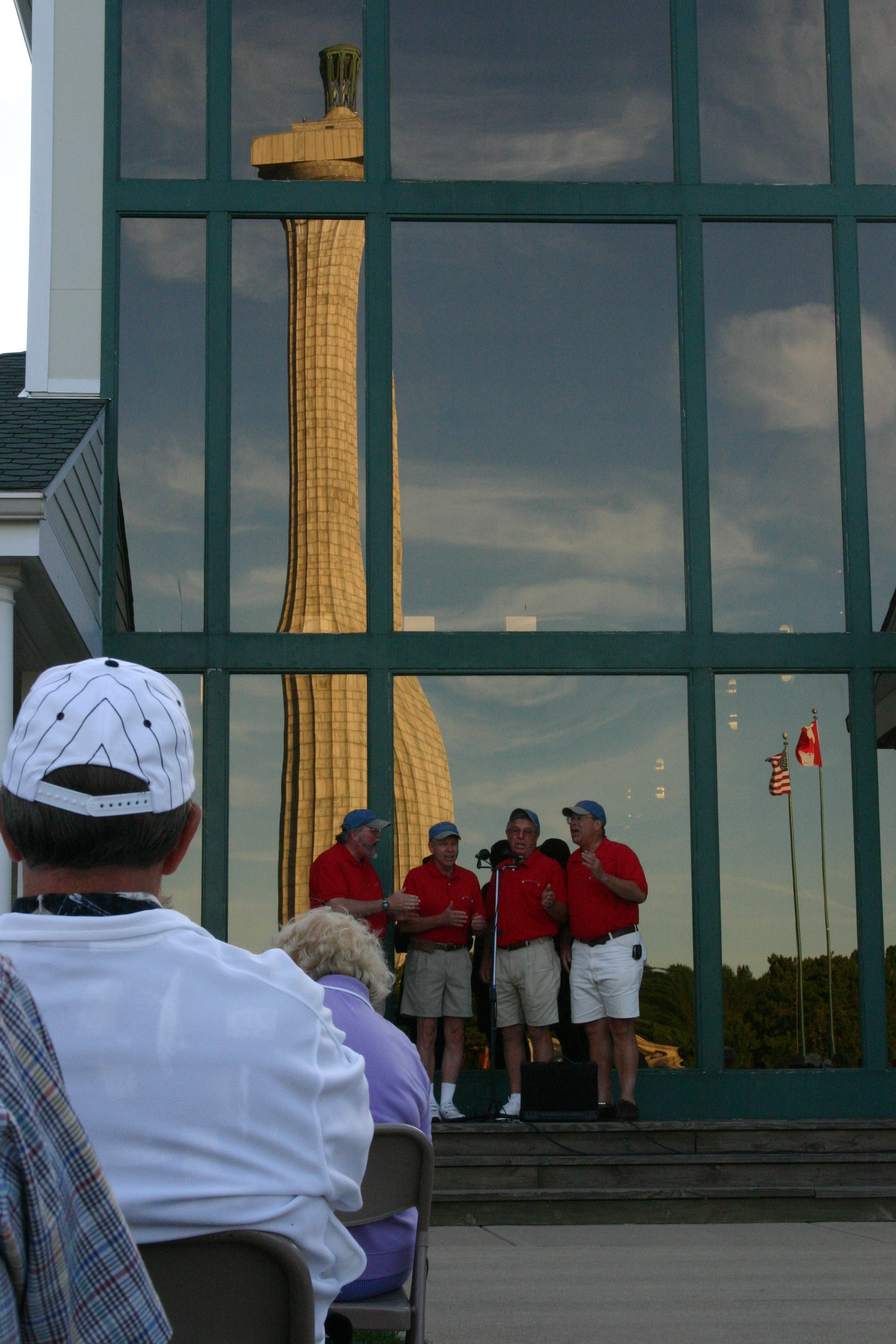 4 Men in red shirts singing in front of a window that reflect a tall stone monument