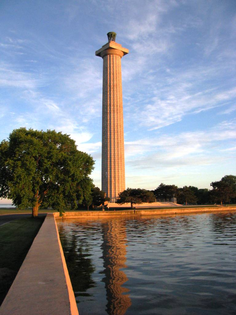 A seawall running in front of 352 foot tall stone memorial. Water reflects the stone memorial.