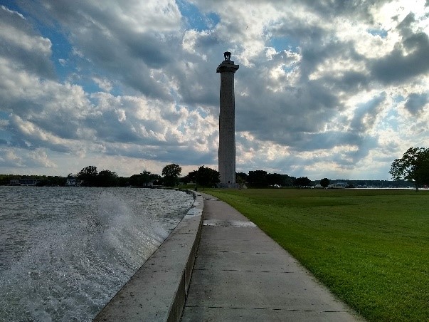 Wave breaking along crumbling seawall. Wave breaks along a crumbling seawall with sidewalk opposite of the water. Sidewalk and seawall is center and curves to the left. Right center is tall stone memorial and grassy lawn.