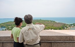 Two visitors stand on observation deck with a stone wall just over waist high They are looking out over large body of water with islands.