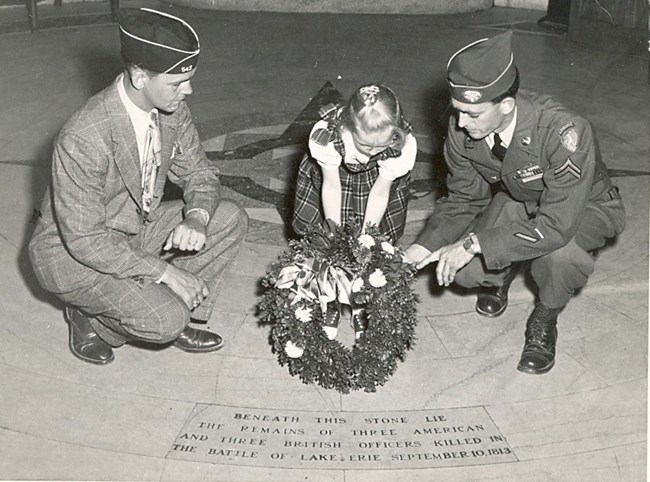 Black and white image of 3 people kneeling to place a wreath near a stone that reads beneath this stone lay the  remains of 6 officers killed in the Battle of Lake Erie.