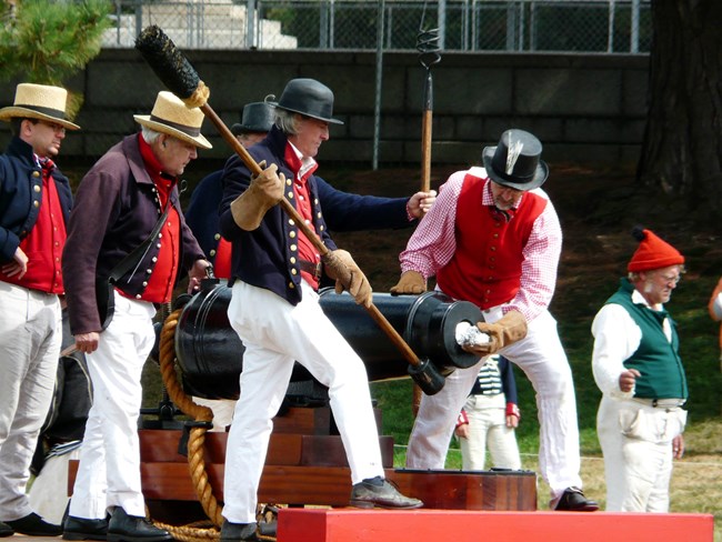Five people dressed as War of 1812 sailors, blue wool jackets, red vests, white pants, with straw or black tarred hats load a large naval cannon.