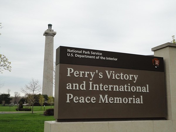 Perry's Victory and International Peace Memorial Sign with Memorial in background. Sign says Perry's Victory and International Peace Memorial and the Memorial is a Greek Doric column made of stone.
