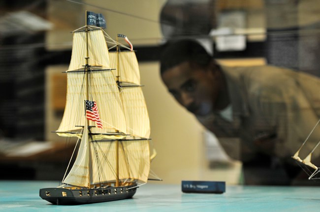 A visitor wearing a Navy uniform peers through a glass case at a ship model