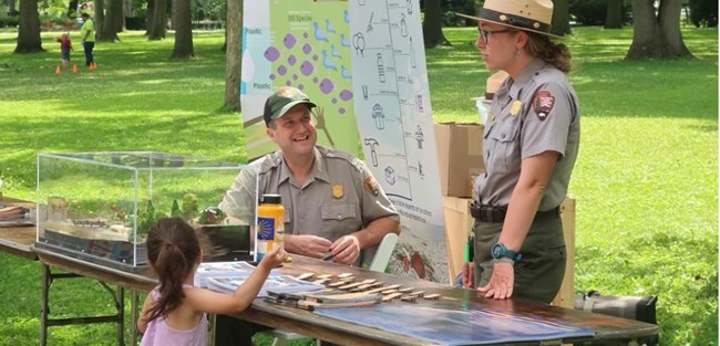 Two park rangers, one seating other standing one one side of a table full of exhibits talk with a young visitor on the other side of the table.